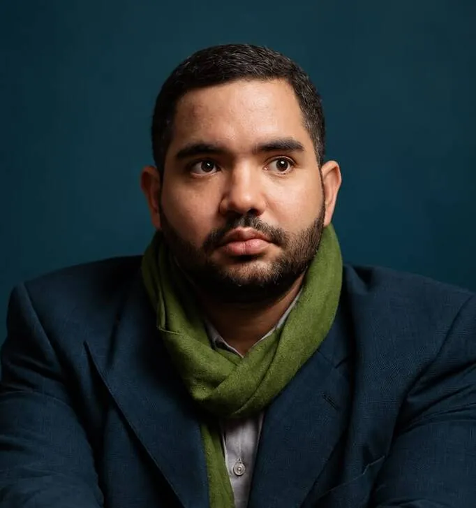 Retrato do Dr. Vitor Piva Moraes, homem de barba, com blazer azul-marinho e cachecol verde, em frente a um fundo azul-petróleo.